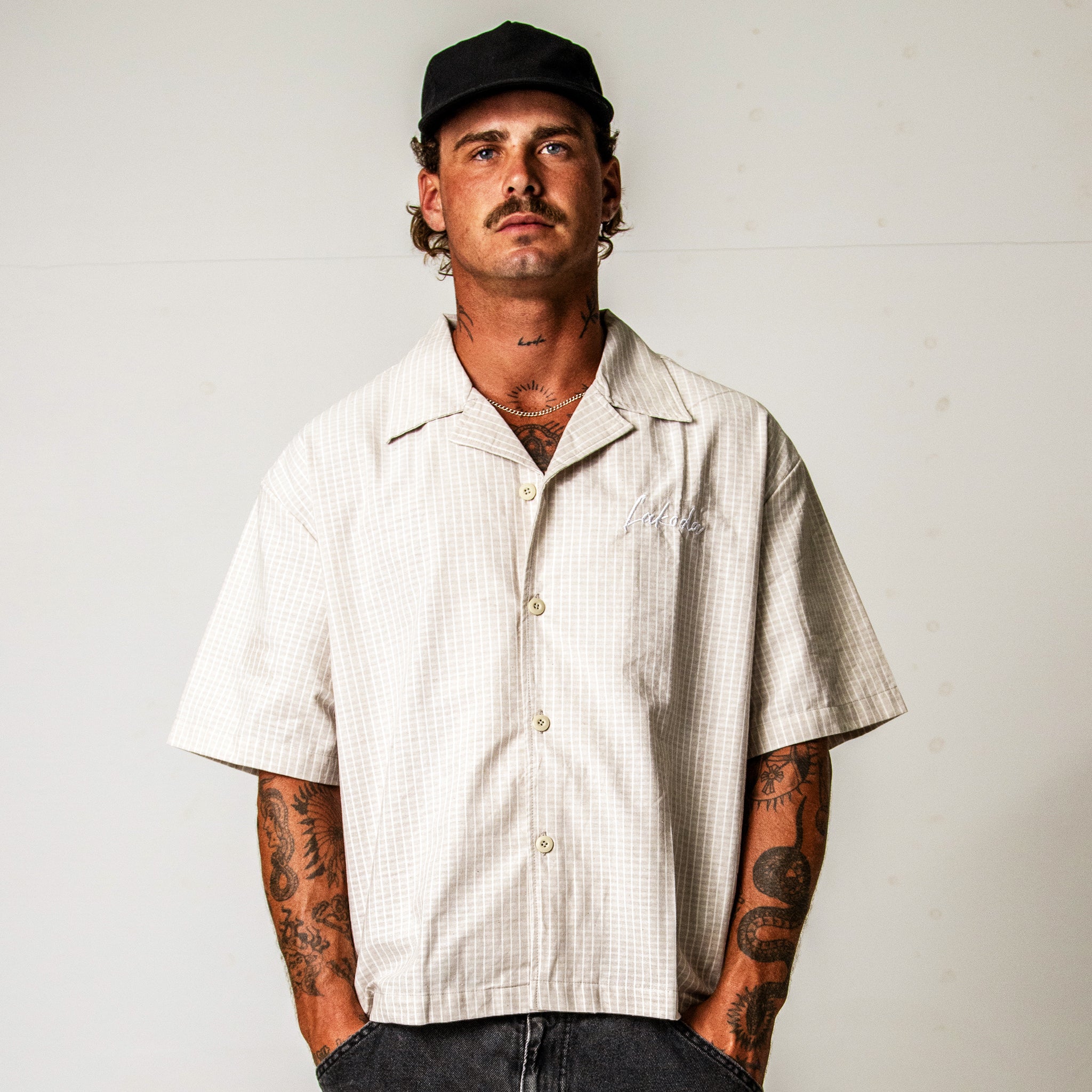 Man wearing a beige short-sleeve shirt and black cap against a plain background